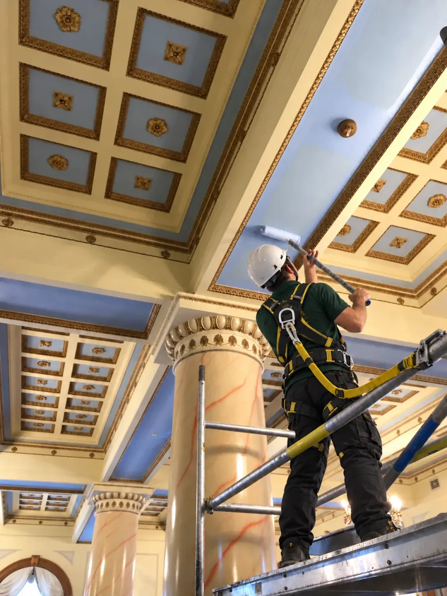 Painter in a green polo, white helmet and safety harness on a scaffold, rolling blue paint onto a coffered ceiling with gilded mouldings beside a marble column
