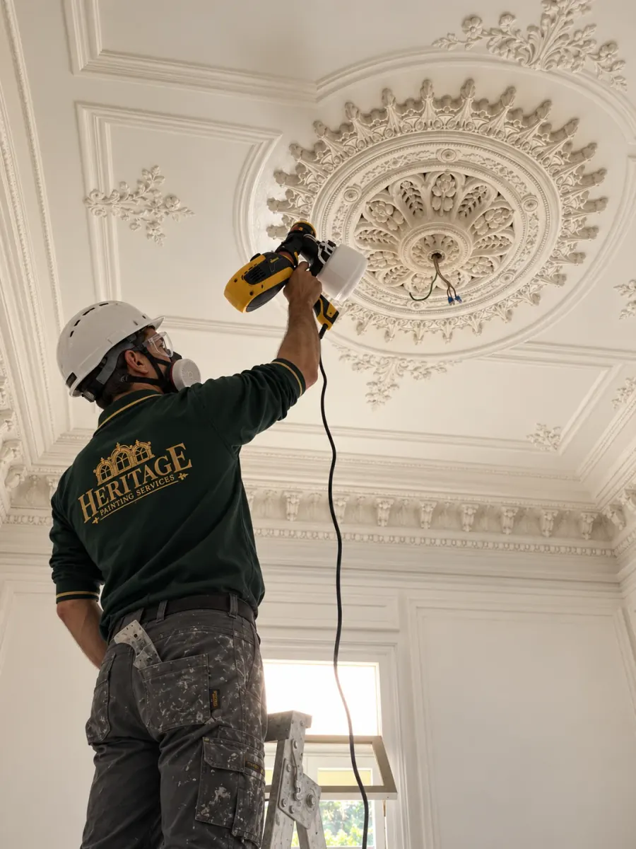 Heritage Painting Services painter in a green polo, helmet and respirator holding a spray gun up to an ornate white plaster ceiling rose, standing on a ladder