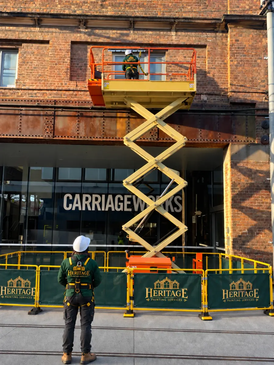 Heritage Painting Services crew member in a green jacket, harness and white helmet watching a colleague on a scissor lift, with green Heritage barriers around the lift in front of a heritage brick building